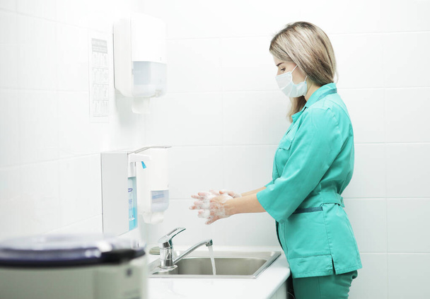 Une femme médecin portant un masque protecteur et un uniforme médical se lave soigneusement les mains à l'hôpital. Mains savonnées en mousse - Photo, image