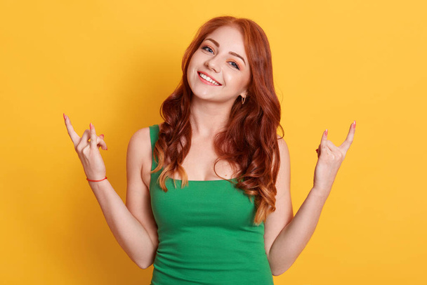 Emotional female makes rock n roll gesture, enjoys cool music at party, demonstrates hand gesture, dressed green outfit, models over yellow background. - Foto, Bild
