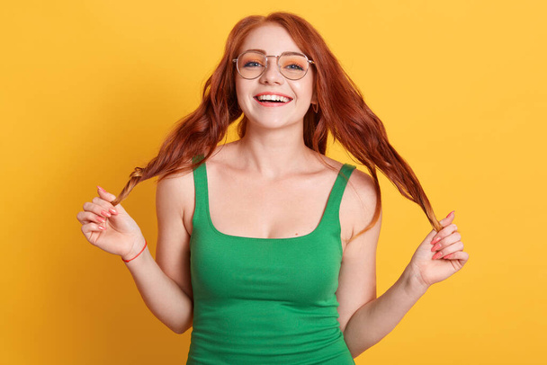 Portraits of charming red-haired girl posing with wonderful mood and lovely smile, touching her wavy beautiful hair, standing against yellow wall. - Fotografie, Obrázek
