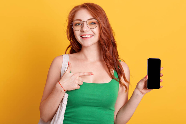 Young beautiful woman showing to camera blank screen of smart phone isolated over yellow background, being in good mood, red haired female wearing green dress. - Φωτογραφία, εικόνα