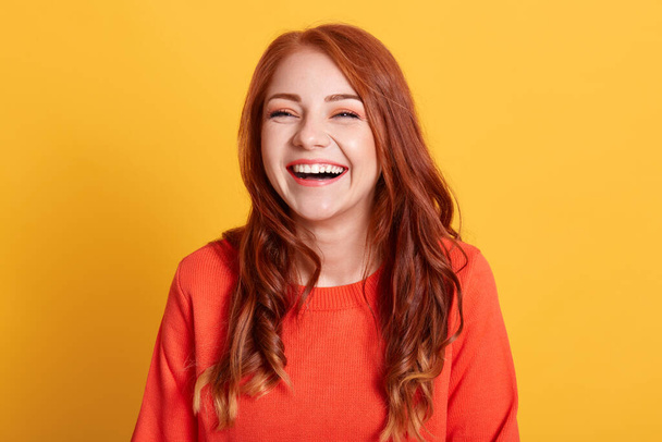 Молодая смеющаяся женщина на жёлтом фоне, выглядит счастливой, носит оранжевый свитер, выражает позитивные эмоции, имеет рыжие волосы, находясь в хорошем настроении. - Фото, изображение