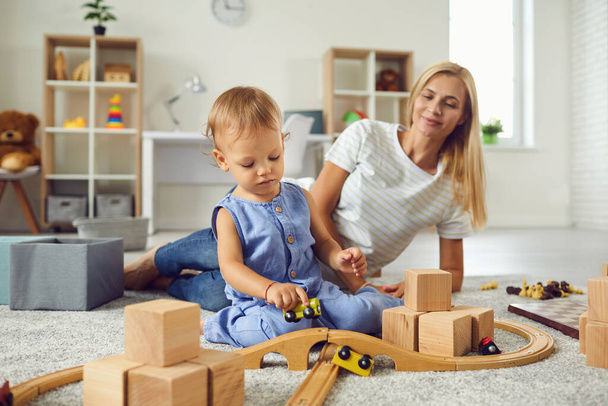 Młoda opiekunka i małe dziecko bawią się drewnianymi klockami w przytulnym pokoju dziecięcym - Zdjęcie, obraz
