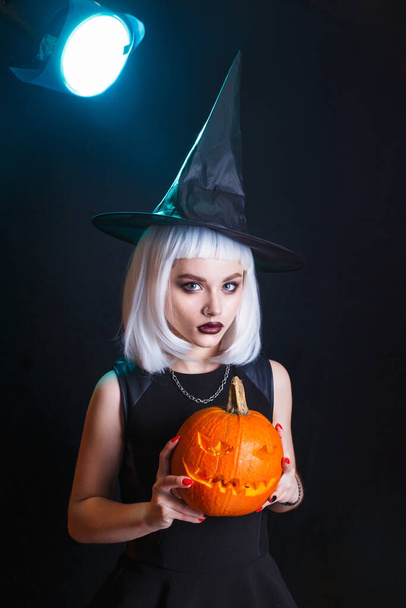 Beautiful young woman in witches hat and costume holding pumpkin on black background - Foto, imagen