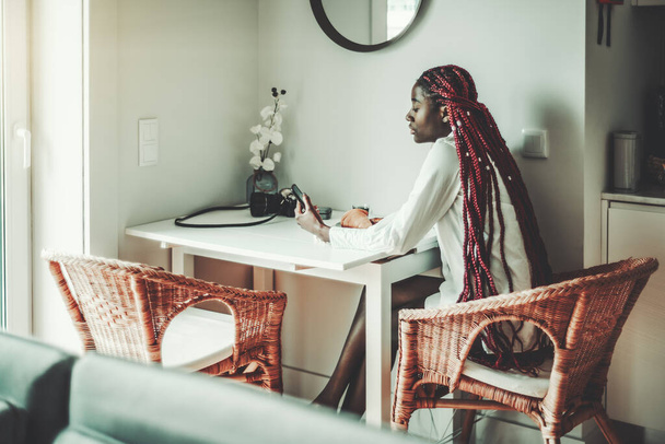 Una encantadora joven hembra negra pensativa con largas trenzas de color rojo oscuro está sentada en un sillón de madera de mimbre en la mesa de la cocina por la mañana durante el desayuno y usando su teléfono inteligente. - Foto, imagen
