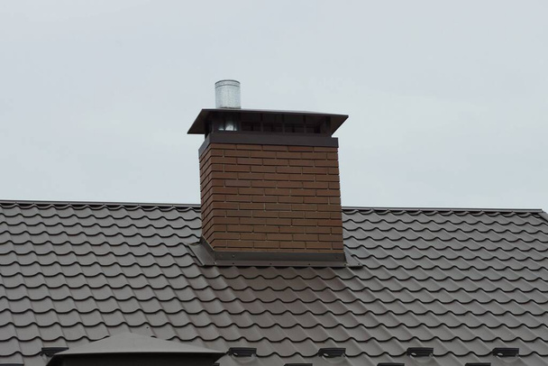 een bakstenen schoorsteen op het bruine betegelde dak van een particulier huis tegen de grijze lucht - Foto, afbeelding