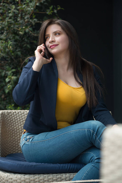 Белая латиноамериканка с длинными волосами и светлыми глазами, сидящая улыбающаяся и разговаривающая по телефону, в желтой футболке, джинсах и свитере, фото с дневным светом - Фото, изображение