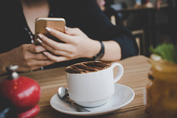 Frau tippt SMS auf Smartphone in einem Café. Zugeschnittenes Bild einer jungen Frau, die mit ihrem Handy an einem Tisch sitzt und einen Kaffee trinkt - Foto, Bild