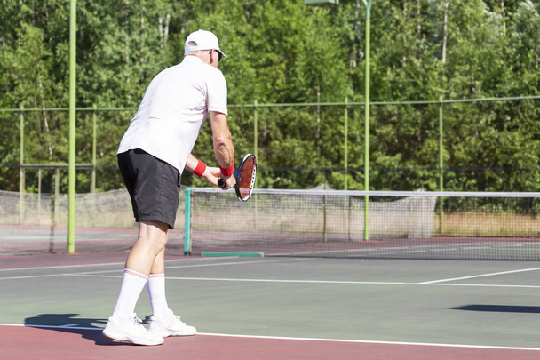 Пожилой человек в хорошей физической форме играет в теннис на открытом воздухе в солнечный день - Фото, изображение
