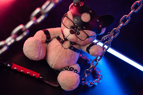 игрушечный медведь, одетый в кожаные ремни безопасности аксессуар для БДСМ игр на темном фоне в неоновом свете - Фото, изображение