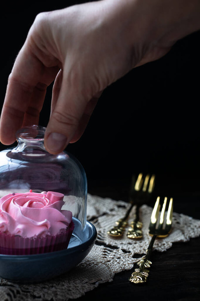 Mão segura uma tampa de vidro e abre um bolo de creme de pé em um guardanapo vintage contra um fundo escuro. - Foto, Imagem