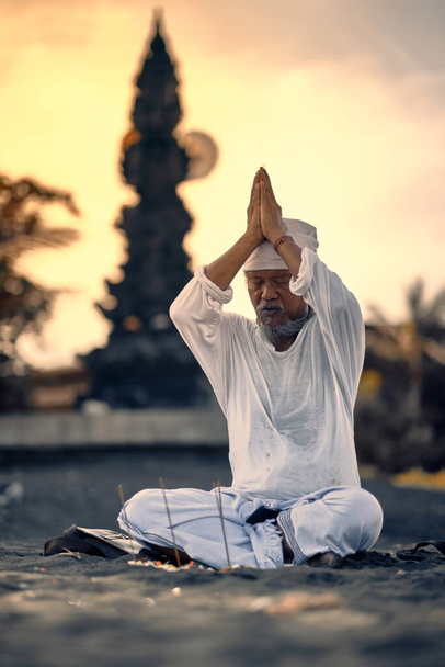 Bali, Indonesia - 10 Dec 2019: Old local balinese man in meditation near the sea. Concept of pray. Adult practice yoga on the Keramas black sand beach at sunset. Rural asian man doing yoga exercise on the beach at sunset. - 写真・画像