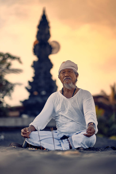 Bali, Indonesia - 10 Dec 2019: Old local balinese man in meditation near the sea. Concept of pray. Adult practice yoga on the Keramas black sand beach at sunset. Rural asian man doing yoga exercise on the beach at sunset. - Foto, Bild