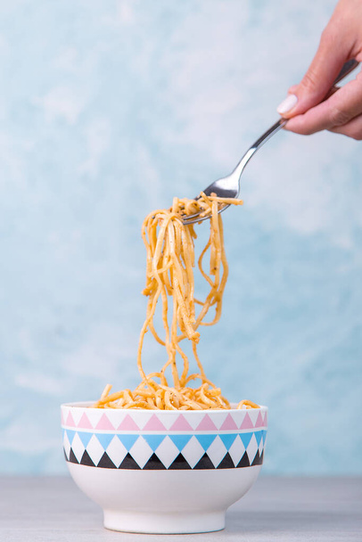 Noedels met saus in gekleurde kom, hand houdt een vork pasta opknoping, eetlust spaghetti op een blauwe achtergrond. - Foto, afbeelding