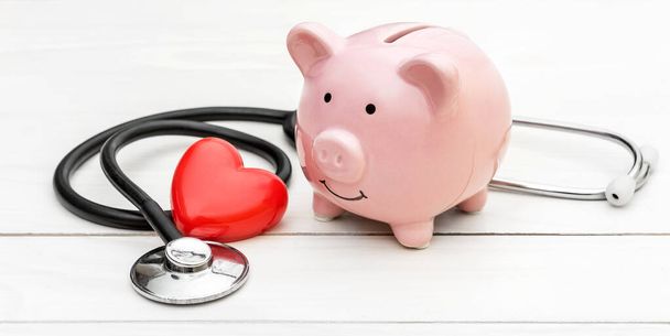 Piggy bank with stethoscope and red heart on white wooden table. - Fotoğraf, Görsel