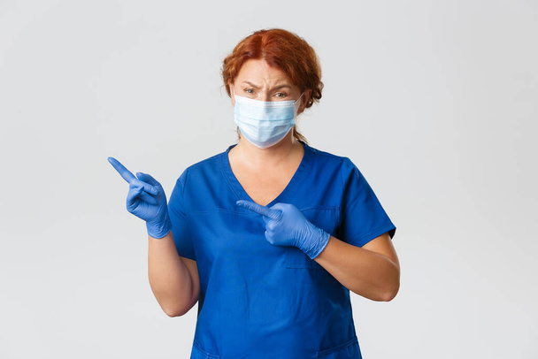 Medical workers, covid-19 pandemic, coronavirus concept. Skeptical and unamused redhead female doctor, physician looking with judgement, pointing upper left corner something unimpressive, wear mask - Fotó, kép