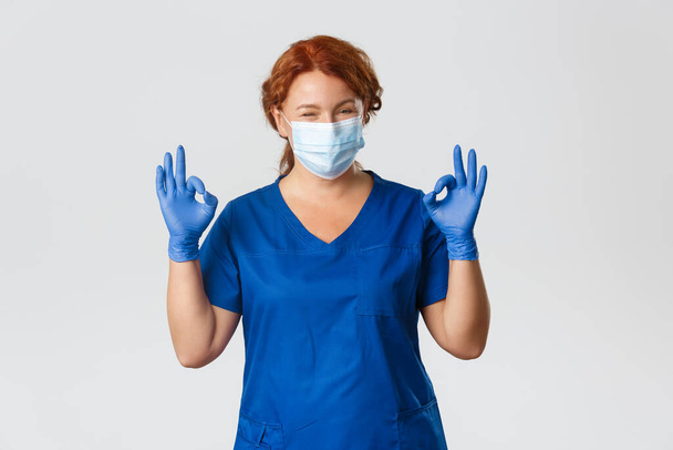 Medical workers, covid-19 pandemic, coronavirus concept. Confident smiling redhead doctor, female nurse in medical mask, gloves, showing okay gesture, guarantee safe and quality checkup at clinic - Photo, Image