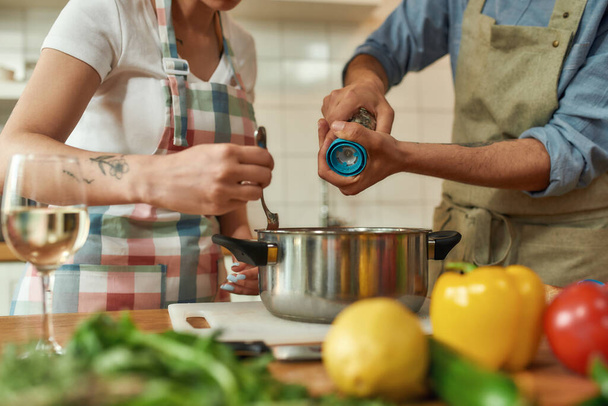 Cropped shot of man adding pepper, spice to the soup while woman stirring it with a spoon. Couple preparing a meal together in the kitchen. Cooking at home, Italian cuisine - Foto, imagen