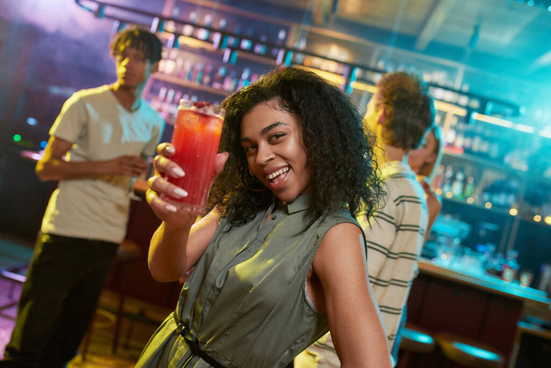 Attractive mixed race young woman looking at camera while posing with a cocktail in her hand and friends chatting, having drinks at the bar counter in the background - Foto, imagen