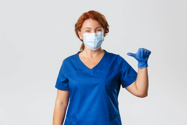 Medizinisches Personal, Covid-19-Pandemie, Coronavirus-Konzept. Angenehme lächelnde Krankenschwester, professionelle Ärztin mit medizinischer Maske, Handschuhe und Peeling, die auf sich selbst zeigt, echter Profi, grauer Hintergrund - Foto, Bild
