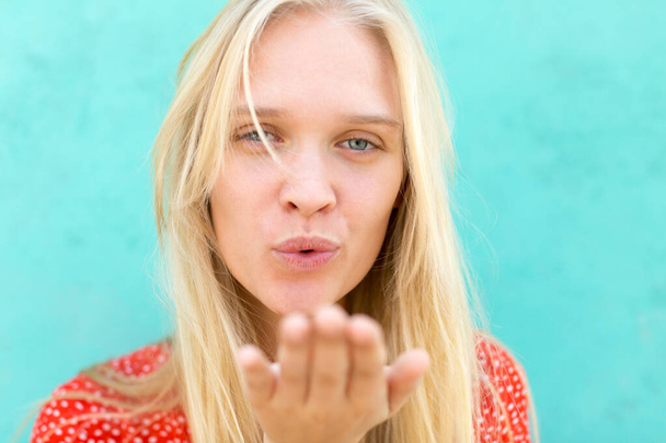 Closeup of pretty blonde girl blowing a kiss to the camera isolated against a turquoise background. - Photo, Image