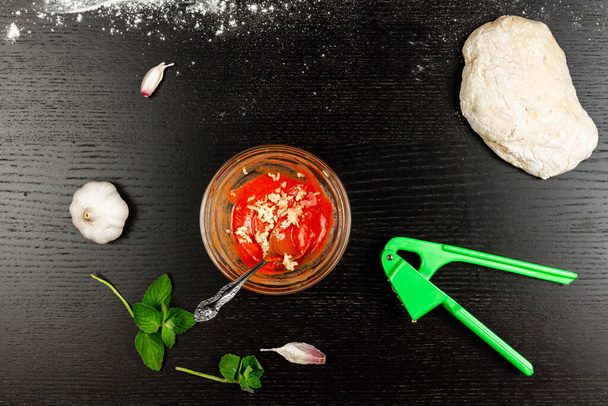 těsto na přípravu pizzy, bylinek, česneku a rajčatové pasty s kořením leží na černém dřevěném stole - Fotografie, Obrázek