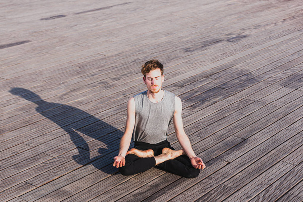 Young red-haired man practicing the lotus yoga posture, Padmasana, outdoors on the wooden floor - Photo, Image