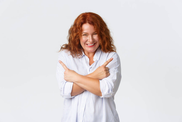 Silly and happy middle-aged redhead woman giggle and and smiling, pointing fingers sideways to show left and right promo offers, found two amazing choices, standing white background - Photo, image