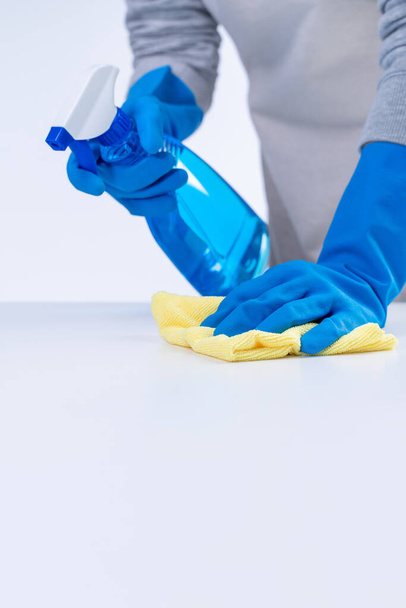 Fiatal nő házvezetőnő kötényben takarít, letörli az asztal felületét kék kesztyűvel, nedves sárga ronggyal, permetező üveg tisztító, closeup design koncepció. - Fotó, kép