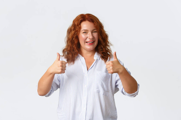 Хорошая работа, хороший жест. Портрет довольной улыбчивой рыжей женщины средних лет поощряет вас, делает комплименты, показывая большие пальцы вверх в одобрении, как продукт, гарантировать качество - Фото, изображение