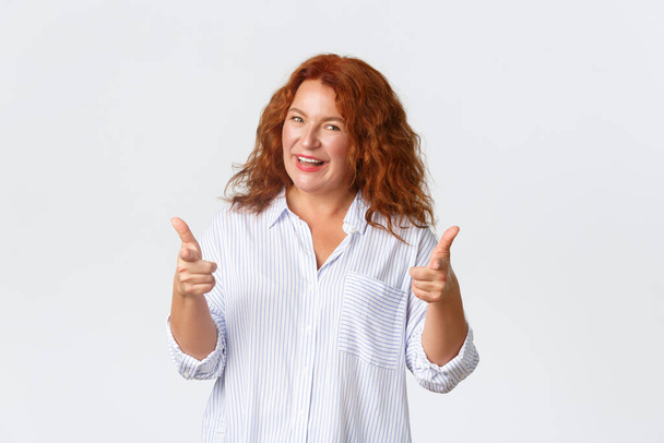 Ritratto di imprenditrice di mezza età, donna adulta con i capelli rossi che fa complimenti, incoraggia a fare qualcosa, puntando il dito contro la fotocamera e sorridente ottimismo, sfondo bianco - Foto, immagini