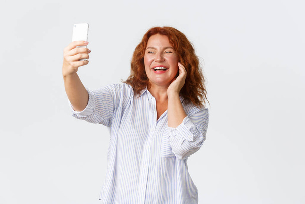 Portré gyönyörű, boldog mosolygós középkorú nő piros hajjal szelfit készít az okostelefonján, pózol fotó szűrővel, alkalmazás, álló fehér háttér - Fotó, kép