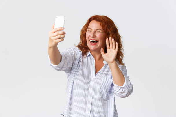 Teknologian ja viestinnän käsite. Ystävällinen ja lähtevä hymyilevä, viehättävä keski-ikäinen punapää nainen puhuu videochatissa, pitelee kännykkää, tervehtii matkapuhelinta ja nauraa - Valokuva, kuva