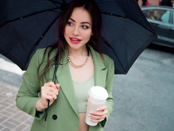 young brunette woman in a green jacket on a walk. Beautiful girl with takeaway coffee - Фото, зображення