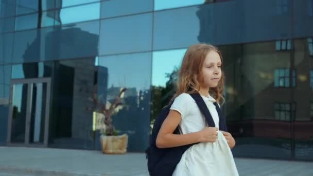 Altın sarısı saçlı şirin bir kız, büyük bir sırt çantası taşıyor, okuldan eve doğru yürüyor, mutlu bir şekilde atlıyor, yol boyunca koşuyor. Öğrenci ilköğretim notları sınıf arkadaşlarına gülümseyerek ders veriyor. - Video, Çekim