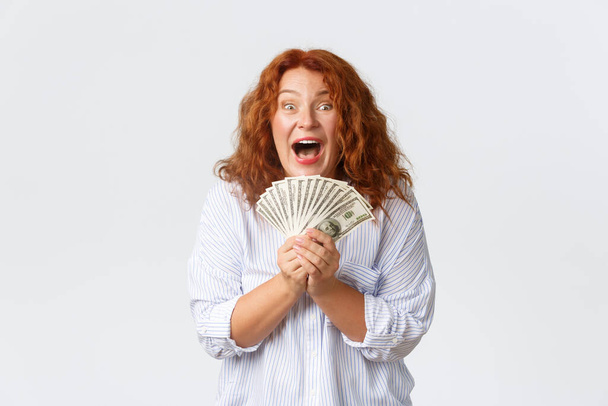 Money, finance and people concept. Triumphing, successful and winning redhead middle-aged woman rejoicing over huge sum of money in her hands, looking thrilled and happy over white background - Photo, Image