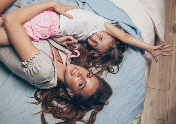Feliz familia cariñosa tumbada relajante disfrutando en dormitorio juntos. Madre y su linda hija preescolar niña en pijama jugando y abrazándose en la cama en la vista superior de la mañana. Feliz día de la madre - Foto, imagen