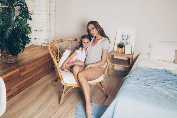 Счастливая любящая семья, отдыхающая в спальне вместе. Мама и ее милая дочка в пижаме обнимаются сидя в плетеном кресле по утрам. С Днем матери! - Фото, изображение