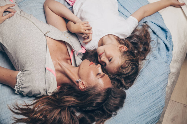 Happy loving family lying relaxing enjoying in bedroom together. Mother and her cute preschool daughter child girl in pajamas playing and hugging in bed in the morning top view. Happy mother's day - Photo, image