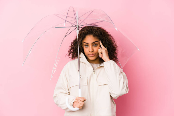 Jonge Afro-Amerikaanse vrouw met een paraplu wijzende tempel met vinger, denken, gericht op een taak. - Foto, afbeelding