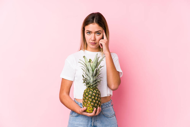 Jonge blanke vrouw met een ananas die zijn tempel met de vinger wijst, denkend, gefocust op een taak. - Foto, afbeelding