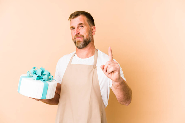 Senior holding a cake isolated on beige background showing number one with finger. - Photo, Image