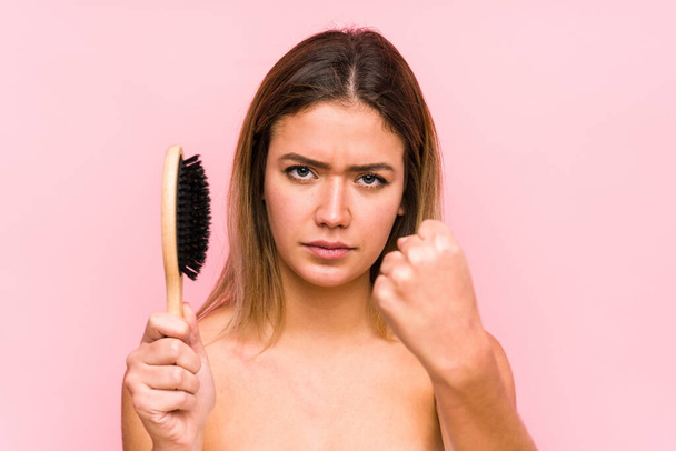 Mulher branca jovem segurando uma escova de cabelo isolado mostrando punho para câmera, expressão facial agressiva. - Foto, Imagem