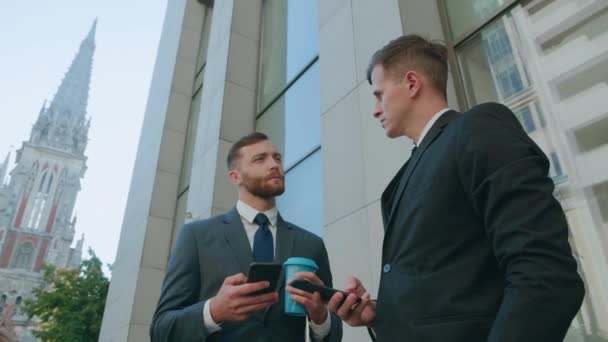 Two male employees interacting with each other in downtown business district. Confident businessmen using smartphone discussing planning computer business project brainstorm in corporate teamwork - Footage, Video