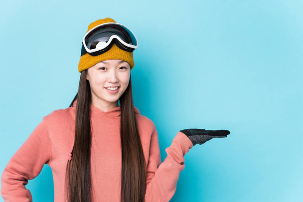 Jeune Chinoise portant des vêtements de ski isolés montrant un espace de copie sur une paume et tenant une autre main à la taille. - Photo, image