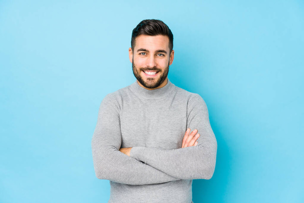 Young caucasian man against a blue background isolated who feels confident, crossing arms with determination. - Photo, Image