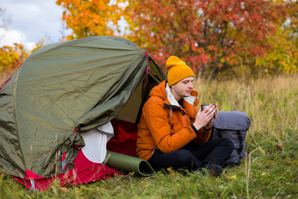 旅行、トレッキング、ハイキングのコンセプト-秋の森の中の緑のテントの近くで茶やコーヒーを飲む若いハンサムな男 - 写真・画像