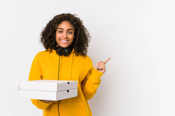 Mladá africká americká studentka držící pizzy s úsměvem a ukazující stranou, ukazující něco na prázdném místě. - Fotografie, Obrázek