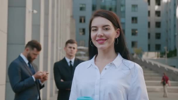 Portrait d'une jeune femme d'affaires prospère à l'extérieur près d'un immeuble de bureaux moderne. Belle employée brune souriant à la caméra sur fond de gens d'affaires. Classique habillé. - Séquence, vidéo