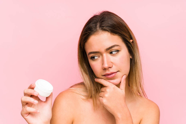 Young caucasian woman holding a moisturizer isolated looking sideways with doubtful and skeptical expression. - Photo, image