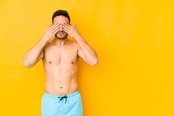 Young caucasian man with swimsuit isolated on yellow background Young caucasian man with trafraid covering eyes with hands. - Photo, Image